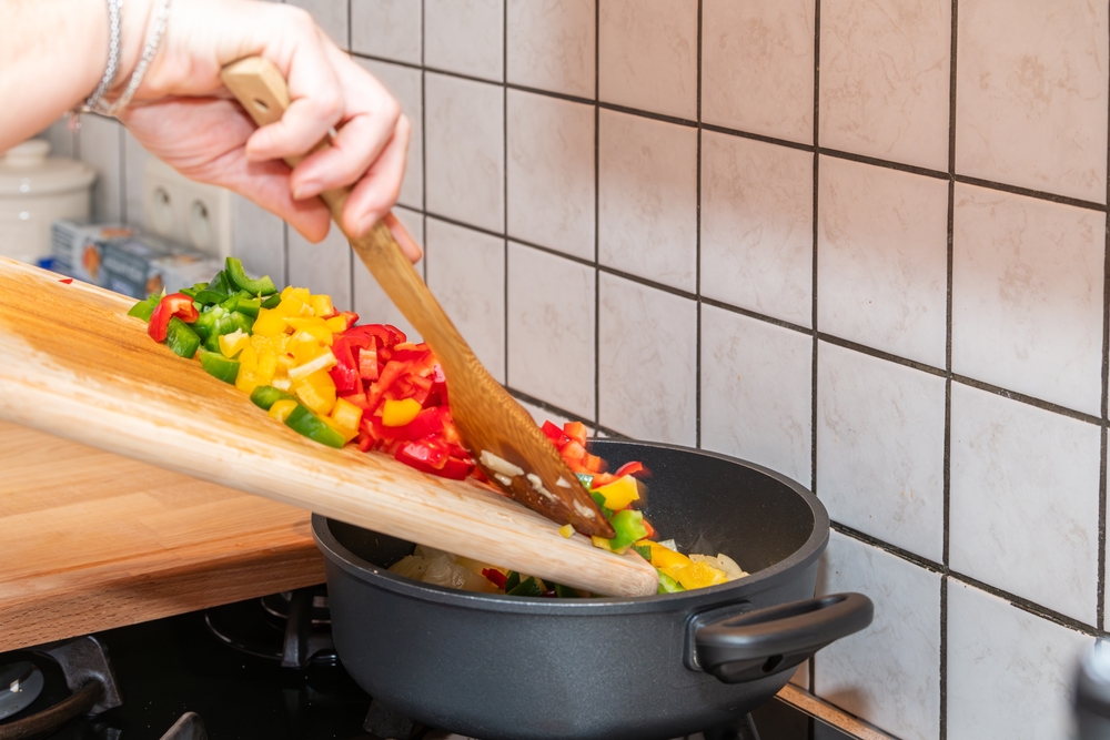 An image showing diced red, yellow and green peppers being scooped into a frying pan from a wooden chopping board.