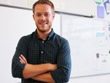 An image of a male teacher with his arms folded, in front of a whiteboard