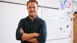 An image of a male teacher with his arms folded, in front of a whiteboard