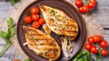 Two grilled chicken breasts on a black plate, accompanied by grilled tomatoes