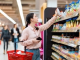 A women looking at a product in the supermarket, whilst carrying a red shopping basket and her phone