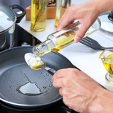 A pair of elderly hands pouring oil into a frying pan, using a spoon to measure the correct amount.