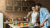 A husband and wife tasting food from a spoon in their kitchen. The counter is covered with colourful vegetables, such as peppers.