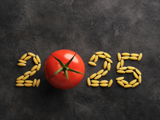 A chalkboard background with the word '2025' written on it. The letters are formed from pasta, except the zero, which is replaced by a tomato